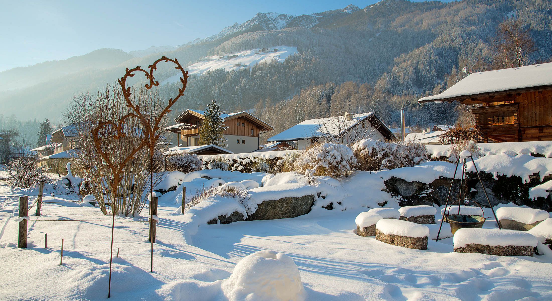 Romantischer verschneiter Garten mit Herzskulptur, Feuerstelle und Bergblick im Winterlicht