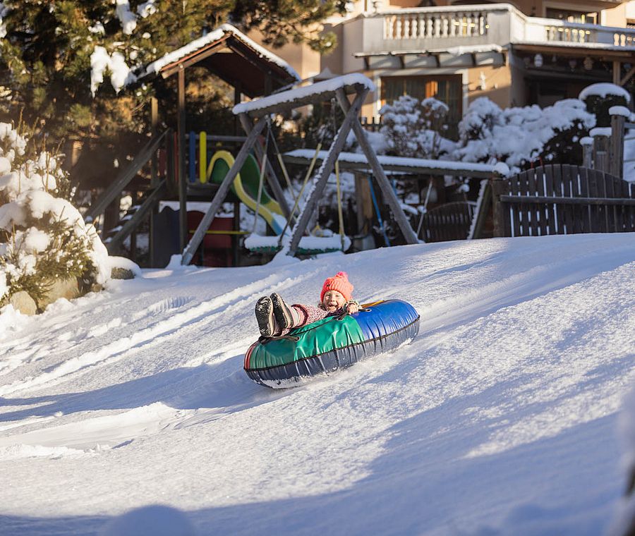 Kind rutscht lachend mit Snowtube eine verschneite Rodelbahn im Hotelgarten hinunter