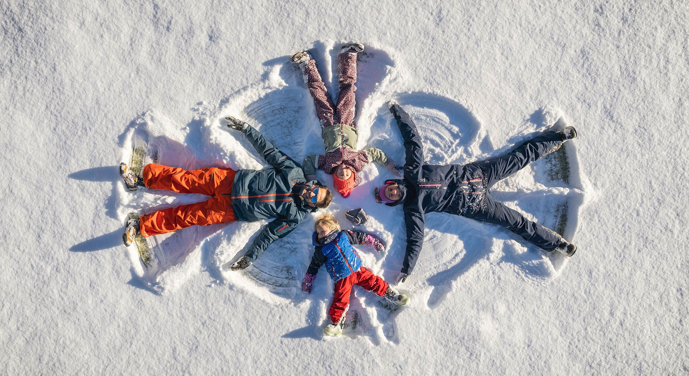 Familie liegt im Schnee und macht gemeinsam Schneeengel – Winterspaß für Groß und Klein
