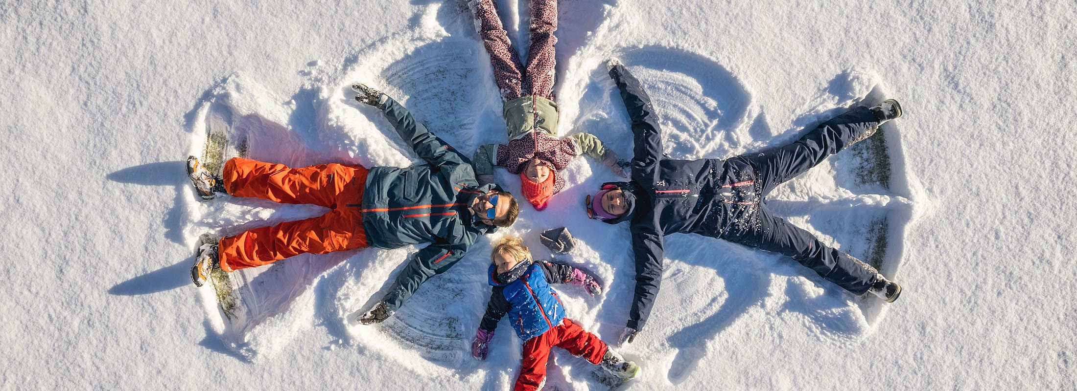 Familie liegt im Schnee und macht gemeinsam Schneeengel – Winterspaß für Groß und Klein
