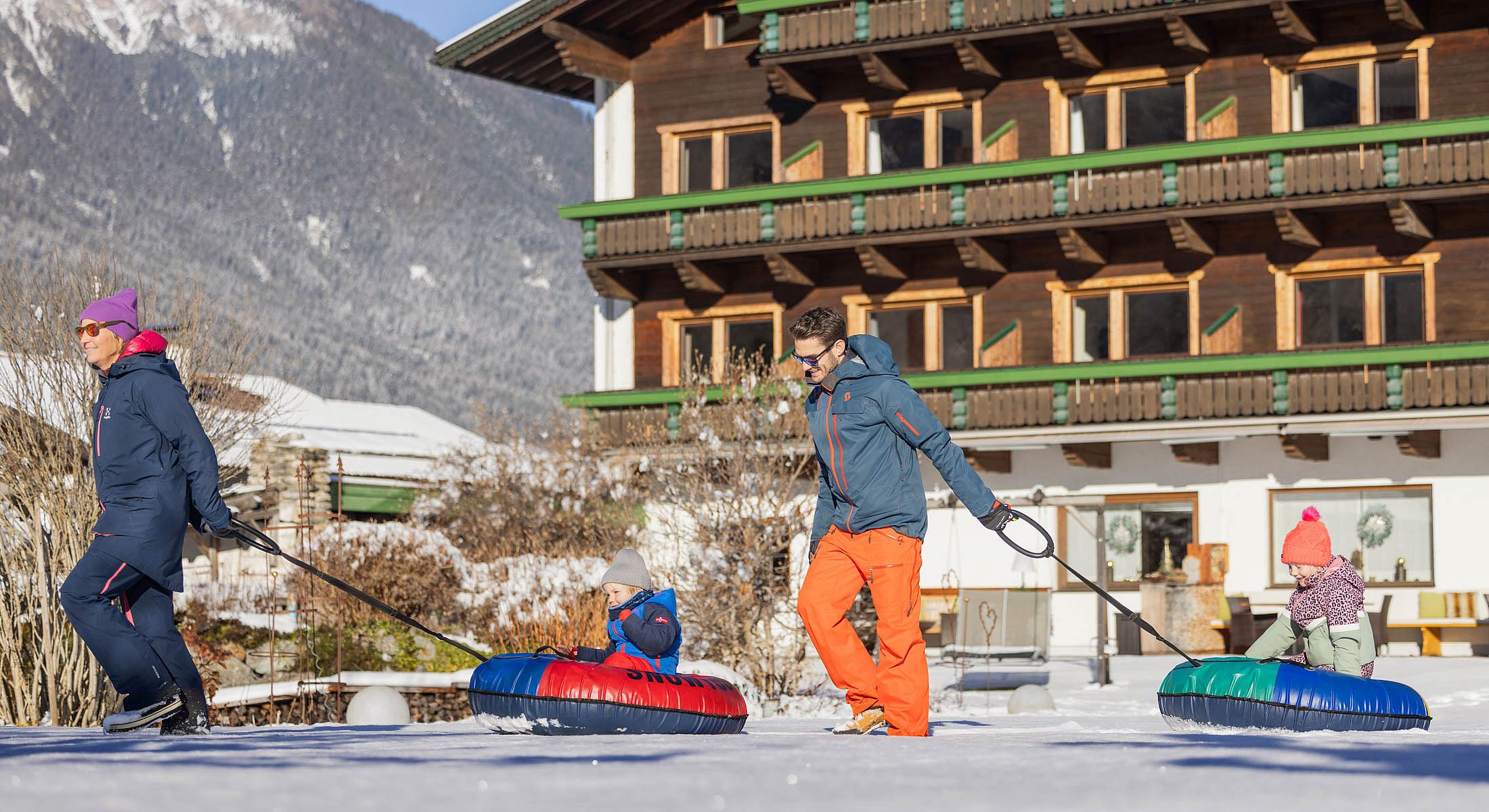 Eltern ziehen ihre Kinder auf bunten Schneereifen durch den verschneiten Garten des Hotels