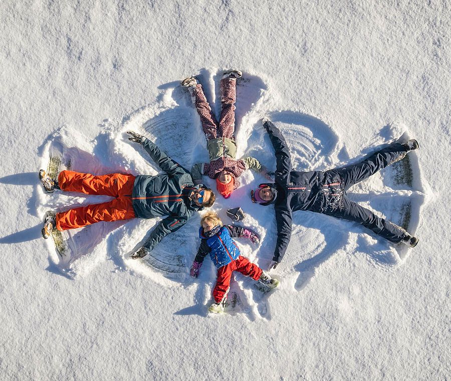Familie macht gemeinsam Schneeengel auf einer schneebedeckten Wiese und lacht dabei