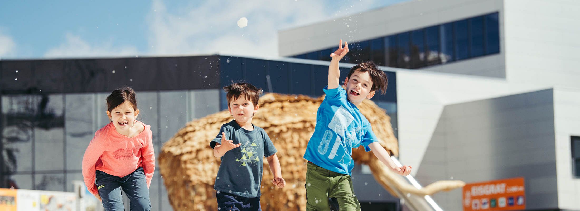 Drei Kinder spielen fröhlich im Schnee vor moderner Architektur mit Kletterspielplatz